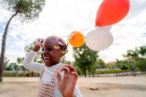 Fille afro-américaine joyeuse avec des tresses dans des vêtements élégants fonctionnant avec des ballons colorés à la main dans le parc en journée — Photo de stock