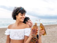 Amando pareja multiétnica de hembras tintineando botellas de cerveza y disfrutando del día de verano en la orilla del mar - foto de stock