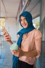 Mulher muçulmana encantada no hijab e com café para ir navegar telefone celular enquanto está na rua da cidade e olhando para a câmera — Fotografia de Stock