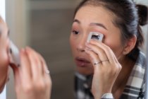 Очаровательная сконцентрированная азиатка, использующая бигуди на ресницах, делая макияж перед зеркалом дома — стоковое фото