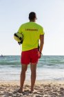 Vue arrière complète du corps du sauveteur masculin anonyme en short et t-shirt et maintien de la sécurité sur le bord de mer sablonneux — Photo de stock