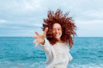 Приємна жінка з довгою імбирною кучерявою зачіскою, що досягає руки до камери, стоячи проти махаючого моря — стокове фото