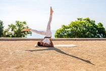 Side view of flexible female in activewear performing Eka Pada Sarvangasana on mat on dry ground during yoga session in park against green trees and cloudless blue sky in sunlight — Stock Photo