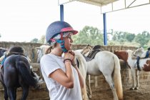Teen girl in casual clothes putting on helmet while standing near horses with saddles in farmyard in stable in daytime — Stock Photo