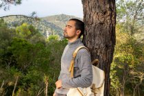 Side view of peaceful young ethnic male hiker in casual clothes and rucksack leaning on tree trunk with closed eyes and enjoying fresh air of green forest in mountainous valley — Stock Photo