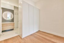 Interior of modern spacious bedroom with white wardrobe placed near door of private bathroom with illuminated oval mirror and wooden furniture — Stock Photo