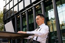 Молодий етнічний чоловік-підприємець, який серфінгує Інтернет на ноутбуці, сидячи за міським столом кафетерії — стокове фото