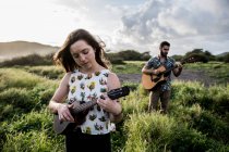 Femme calme en vêtements décontractés jouant ukulele et debout dans le champ vert contre un ami concentré jouer de la guitare dans la nature dans la journée ensoleillée — Photo de stock
