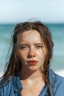 Mujer en camisa mojada y con el pelo mojado de pie mirando a la cámara en la playa cerca del mar mientras disfruta del día de verano - foto de stock