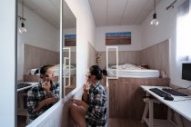 Seitenansicht einer jungen ethnischen Frau, die in der Nähe eines Spiegels sitzt, während sie Puder im hellen Schlafzimmer mit Bett und Schreibtisch mit Computer aufträgt — Stockfoto