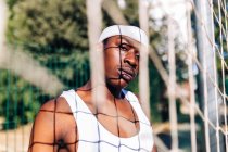 Ernsthafter afroamerikanischer Sportler in weißer Aktivkleidung steht im Sommer in der Nähe eines Netzes auf einem Spielplatz und blickt in die Kamera — Stockfoto