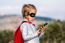 Side view of self assured girl in eye mask superhero costume with cape browsing on cellphone — Stock Photo