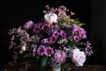 Buquê de peônias coloridas frescas e crisântemos em vaso branco colocado sobre mesa de madeira em quarto escuro — Fotografia de Stock