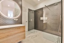 Просторная стеклянная душевая кабина и светящееся овальное зеркало, висящее на стене над раковиной, в просторной современной ванной комнате с мраморным полом — стоковое фото