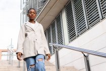 Dal basso di donna afro-americana fiduciosa in abito alla moda in piedi sulla strada vicino edificio con ringhiera in metallo in città mentre si cammina giù per le scale — Foto stock