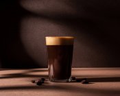 Vorderseite einer schwarzen Kaffeetasse mit einer Schicht Schaum, die neben ein paar Kaffeebohnen auf einem braunen Tisch und einer Wand mit dunklen Schatten steht — Stockfoto