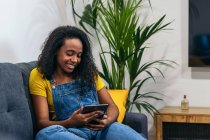 Mulher afro-americana positiva em roupa jeans sentado no sofá e sorrindo enquanto navega tablet em casa — Fotografia de Stock