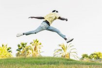 Von unten springt ein junger afroamerikanischer Mann in lässigem Outfit mit weit auseinander stehenden Beinen und erhobenen Händen im Park — Stockfoto