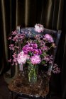 Strauß frischer bunter Pfingstrosen und Chrysanthemen in Glasvase auf verwittertem Holzstuhl in der Nähe von Vorhängen im hellen Raum — Stockfoto