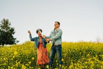 Jovem feliz em roupas casuais abrindo garrafa de champanhe durante encontro romântico com a alegre namorada afro-americana no prado florescente no campo — Fotografia de Stock