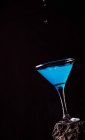 Drops of blue Lagoon cocktail pouring in crystal elegant glass placed on rough surface against black background — Stock Photo
