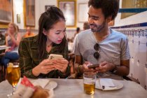 Счастливая молодая многорасовая пара в повседневной одежде с помощью смартфонов, сидя вместе за столом с бокалами пива в современном ресторане — стоковое фото