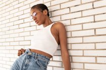 Confident African American female with short hair in stylish outfit with trendy eyeglasses standing on street near white brick wall — Stock Photo