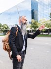 Side view of serious bearded male in formal wear standing near road while hailing taxi on street with modern buildings — Stock Photo