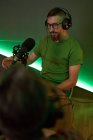 Young male radio host in casual clothes and headphones sitting at table with microphone and communicating with crop anonymous colleague during podcast recording in studio — Stock Photo