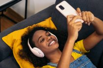 De cima de jovem afro-americano feminino com cabelo encaracolado em fones de ouvido e roupas casuais deitado no sofá cinza confortável e tomando selfie no smartphone na sala de luz em casa — Fotografia de Stock
