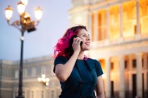 Вид сбоку жизнерадостной женщины с розовыми волосами, которая звонит по телефону, стоя на улице с фонарем возле классического здания в городе — стоковое фото