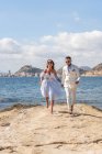 Corpo inteiro de alegre casal de casamento descalço correndo na costa perto do mar ondulante enquanto desfruta do dia do casamento na natureza ensolarada — Fotografia de Stock