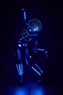 Persona sin rostro en traje brillante contemporáneo de cyborg espacio con iluminación de neón y casco de pie sobre fondo negro en estudio oscuro - foto de stock