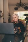 Літня жінка з коротким сірим волоссям сидить на стільці роблячи відеодзвінки через нетбук вдома — стокове фото