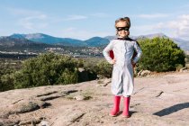 Full body of small girl in superhero costume with hands on waist standing on rocky hill — Stock Photo