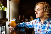 Thoughtful male with curly hair in casual clothes sitting at wooden counter near window in bar and drinking beer in daytime — Stock Photo