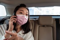 Pasajera étnica con máscara protectora sentada con cinturón de seguridad y dando instrucciones al taxista mientras está en una llamada telefónica - foto de stock