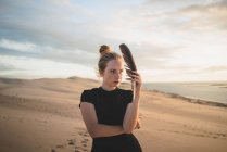 Серйозна жінка з чорним пір'ям у сукні, що стоїть на піщаній дюні, омитій морем під час заходу сонця — стокове фото