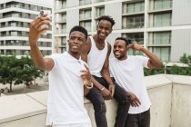 Homem afro-americano positivo com amigos mostrando gesto de vitória enquanto companheiro tirando selfie no celular — Fotografia de Stock