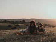 Hippie-Paar mit Sonnenbrille blickt in die Kamera, während es bei Sonnenuntergang auf einer Wiese in der Natur liegt — Stockfoto