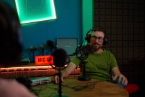 Positif jeune homme barbu millénium dans les lunettes et écouteurs et parler en micro tout en enregistrant podcast en studio sombre — Photo de stock