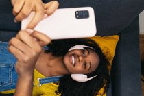 From above of young African American female with curly hair in headphones and casual clothes lying on comfortable gray sofa and taking selfie on smartphone in light room at home — Stock Photo