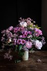Bouquet de pivoines fraîches colorées et de chrysanthèmes dans un vase blanc posé sur une table en bois dans une pièce sombre — Photo de stock