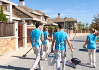 Back view of unrecognizable young cleaning crew in uniform carrying brooms with scoops and buckets while walking on paved street near residential houses on sunny day — Stock Photo