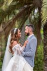 Couple marié dans des tenues de mariage debout sur la passerelle en bois avec rampe tout embrassé et en regardant les uns les autres près des palmiers verts et des plantes dans le jardin en journée d'été — Photo de stock