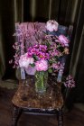 Ramo de peonías frescas y coloridas y crisantemos en jarrón de vidrio colocado en la silla de madera envejecida cerca de las cortinas en la sala de luz - foto de stock