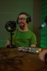 Позитивний молодий бородатий чоловічий тисячоліття в окулярах і навушниках посміхається і розмовляє в мікрофоні під час запису подкасту в темній студії — стокове фото
