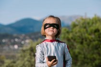 Menina auto-assegurada no traje de super-herói máscara olho com capa de navegação no celular — Fotografia de Stock