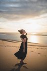 Seriöses Weibchen mit schwarzer Feder trägt Kleid, das auf einer Sanddüne steht, die bei Sonnenuntergang vom Meer gewaschen wird — Stockfoto