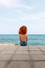 Vista posteriore di femmina irriconoscibile con riccioli di zenzero seduti su argine di pietra e ammirando il paesaggio marino — Foto stock